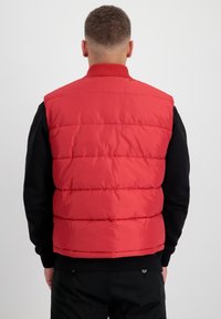 Doudoune rouge avec un motif matelassé, col côtelé et coupe sans manches. Portée par-dessus une chemise noire à manches longues. Texture lisse.
