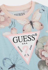 Maglietta azzurro chiaro con un motivo floreale e farfalloso, un'etichetta triangolare rosa con il testo "GUESS" e una finitura lucida.