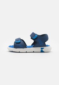 Sandalo blu e navy con cinturini regolabili in Velcro, una soletta blu testurizzata e una suola in gomma bianca con un motivo a zigzag.