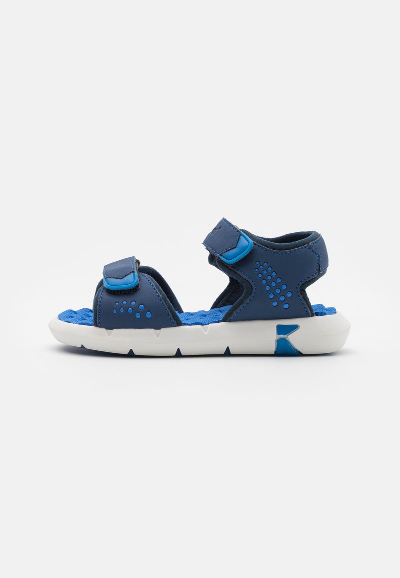 Sandalo blu e navy con cinturini regolabili in Velcro, una soletta blu testurizzata e una suola in gomma bianca con un motivo a zigzag.