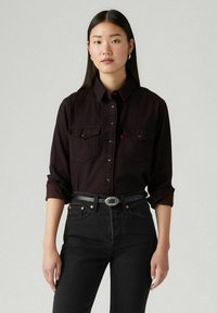 Chemise noire à boutons avec deux poches poitrine, col et manches retroussées. Portée avec un jean noir taille haute et une ceinture texturée avec une boucle ronde.
