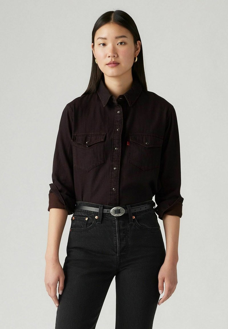 Chemise noire à boutons avec deux poches poitrine, col et manches retroussées. Portée avec un jean noir taille haute et une ceinture texturée avec une boucle ronde.