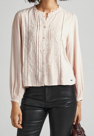 Frau trägt eine hellrosa, langärmelige Bluse mit Knöpfen und bestickten Details, kombiniert mit schwarzer Hose und hält eine bordeauxrote Handtasche.