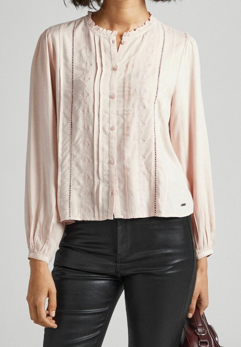 Femme portant un chemisier rose pâle à manches longues avec des boutons et des détails brodés, associé à un pantalon noir et tenant un sac à main bordeaux.