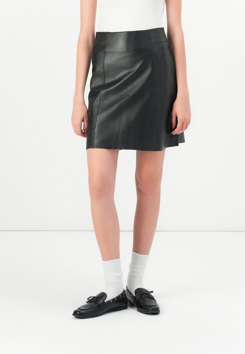 Svart skinn miniskjørt med en tettsittende midje, lett utsvingt hem og synlige sømdetaljer. Brukt med svarte loafers og hvite sokker.