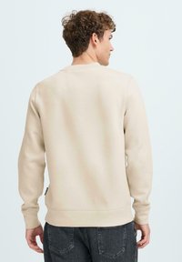 Beige sweatshirt met lange mouwen en ronde hals. Soepele textuur, ribgebreide manchetten en zoom. Draag met donkergrijze jeans, bekeken vanuit de achterkant.