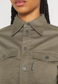 Olivgrön skjorta i mjukt tyg, med spetsig krage, två framfickor och mörka knappar med en svart etikett.