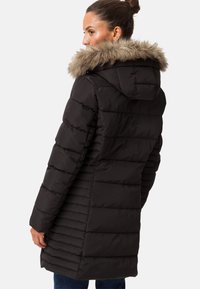 Svart puffjacka med foder i fuskpäls, med kviltad textur, långa ärmar och en medellång längd. Bakifrån visas den figursydda designen.