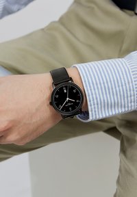 Reloj analógico negro con una carátula redonda, marcadores de hora en contraste blancos, correa de cuero negro y acentos rojos. Lleva puesto en una muñeca.