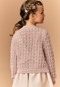 Cardigan tricotat în nuanță roz deschis, cu un model de dantelă deschis, guler rotund și manșete striate. Purta peste o fustă deschisă, cu părul coafat cu flori.