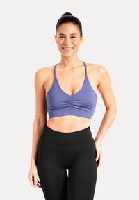 Kvinna med hår i en knut, iförd en lila sport-bh och svarta högmidjade leggings, som ler och poserar mot en enfärgad bakgrund.