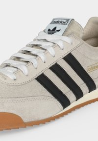 Beiger Adidas-Sneaker mit weißen Schnürsenkeln, schwarzen drei Streifen, Gummisohle und Adidas-Logo auf Zunge und Seite.