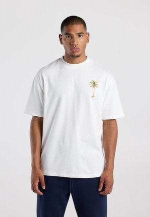 Man met kort haar die een wit T-shirt draagt met een kleine palmboomafbeelding op de borst en donkere broek, staande tegen een effen achtergrond.
