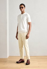 Hvit piké skjorte, lyse beige bukser og mørkebrune lærsko. Modellen har på seg solbriller, står på et tregulv mot en lys bakgrunn.