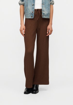 Persoon die een textuurbruine broek met wijde pijpen en trekkoord draagt, zwarte enkellaarsjes, een wit shirt en een lichtblauwe opgerolde spijkerjack.