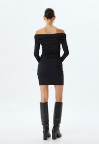 Schwarzes, schulterfreies Kleid mit langen Ärmeln und Raffung, kombiniert mit hohen schwarzen Stiefeln mit Absatz. Glatter, dehnbarer Stoff mit taillierter Silhouette.