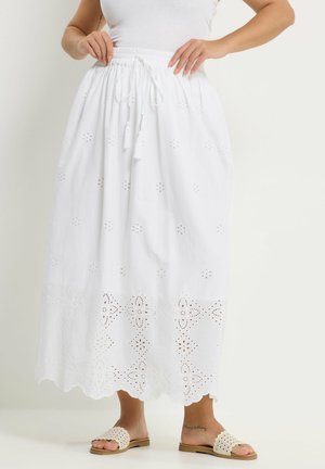 Kobieta ubrana w białą midi spódnicę z ażurowymi kwiatowymi wycięciami i wiązanym w talii sznurkiem z frędzlami, do tego białe szydełkowane sandały wsuwane.