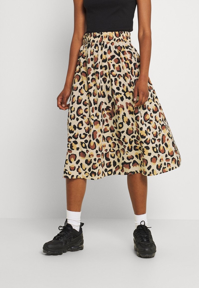 Femme portant une jupe midi à imprimé léopard, des baskets noires, des chaussettes blanches et un haut sans manches noir, debout les jambes légèrement écartées.