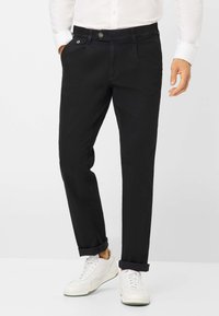 Schwarze, enganliegende Chinos mit glatter Oberfläche, ausgestattet mit einem Knopfverschluss, Taschen und umgeschlagenen Säumen, kombiniert mit weißen Sneakers.