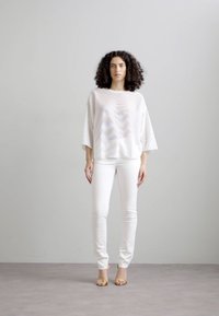 Top alb, lejer, cu un model texturat, cu mâneci scurte, combinat cu blugi skinny albi și sandale aurii, pe un fundal neutru.