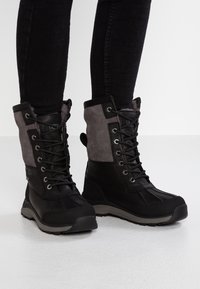 Czarno-szare zimowe botki na sznurówki z futrzanym wnętrzem, noszone z dopasowanymi czarnymi spodniami na białym tle.