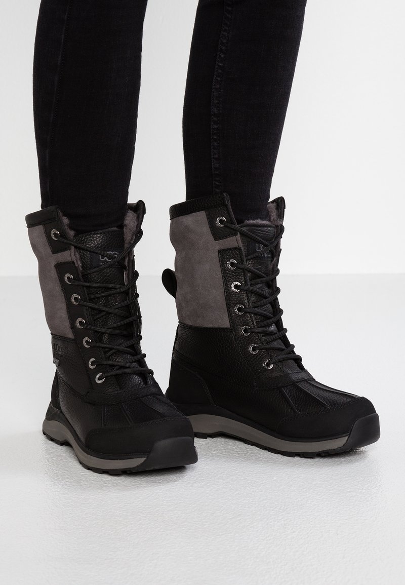 Czarno-szare zimowe botki na sznurówki z futrzanym wnętrzem, noszone z dopasowanymi czarnymi spodniami na białym tle.