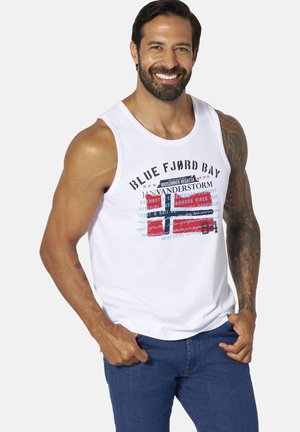 Man with beard wearing white sleeveless shirt with nautical-themed graphic and blue jeans, smiling with hands in pockets.