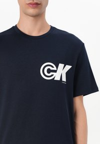 Námornícky modré bavlnené tričko s veľkým bielym grafickým logom na ľavej strane hrude, s okrúhlym výstrihom a krátkymi rukávmi.