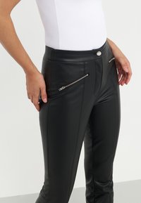 Pantalons en simili cuir noir à coupe slim, avec deux zips sur les côtés, une taille plate et une texture lisse, affichés avec une main sur la hanche.