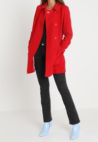 Abrigo de mezcla de lana rojo con cuello de muesca y botones de doble botonadura, combinado con unos vaqueros negros y botas de tac ón brillantes en azul claro.