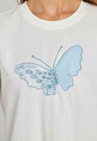 T-shirt bianco in cotone con una farfalla ricamata blu, decorata con perline e dettagli in pizzo, che mette in mostra intricati motivi e texture.