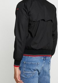 Man i svart bomberjacka med ribbade cuffs och midja, över blå jeans med bakfickor, sedd från baksidan.