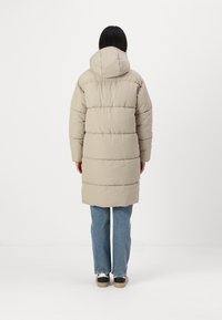 Casaco puffer bege com capuz, apresentando costuras horizontais, design até ao joelho e bolsos frontais. Combinado com jeans de um azul claro e sapatos pretos.