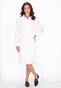 Witte lange mouw jurk met knoopsluiting en kraag, gemaakt van gladde stof, valt net boven de knie, gecombineerd met zwarte loafers.