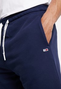 Main dans la poche, portant un pantalon de survêtement bleu marine avec un cordon blanc, étiqueté "Tommy Jeans" et un petit patch logo rouge, blanc et bleu.