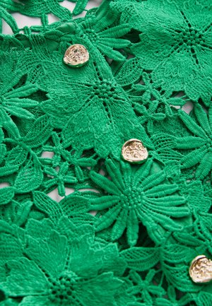 Tissu en dentelle florale verte avec des motifs complexes, des détails texturés et un bouton circulaire doré en accent.