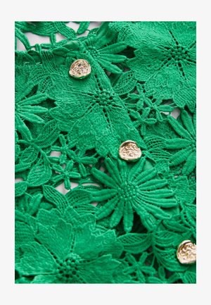 Tessuto in pizzo floreale verde con motivi intricati, dettagli testurizzati e un bottone circolare dorato come accento.