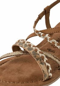 Sandal in suede marrone con cinturini intrecciati, dotato di accenti metallici e decorativi. Suola interna morbida e suola testurizzata per il comfort.
