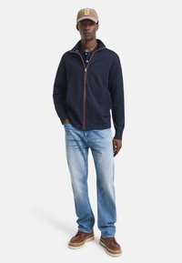 Chaqueta de estilo navy con cremalleras y acentos marrones, combinada con pantalones de mezclilla claros y zapatillas marrones. La chaqueta cuenta con un cuello alto y bolsillos laterales.