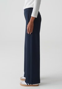 Navy wijde broek met een gladde stof, hoge taille, zijzakken en een getailleerde pasvorm. Gecombineerd met witte en grijze sportschoenen.