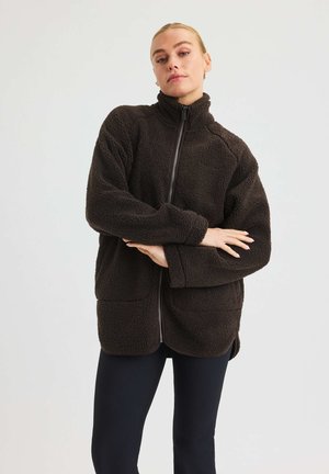 Vrouw in een donkerbruin fleece jack met rits en hoge kraag, staand met gekruiste armen, tegen een effen lichte achtergrond.