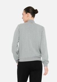 Felpa grigia con colletto alto, polsini e orlo a costine, che presenta una texture liscia. Dettaglio del logo dorato vicino al colletto.