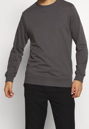 Mann trägt ein schlichtes dunkelgraues Sweatshirt mit Rundhalsausschnitt und langen Ärmeln sowie schwarze Hose, steht mit leicht angewinkelten Armen.