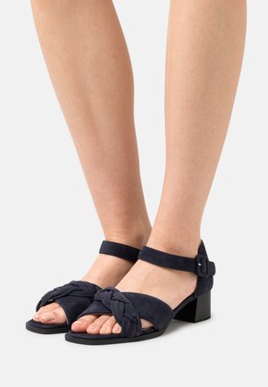 Navy suede sandals with a knotted strap design, open toes, and ankle strap featuring a buckle. Square block heel for stability.