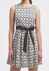 Robe sans manches avec un col rond, présentant un motif noir et blanc, taille cintrée avec une ceinture en satin noir, et jupe évasée.