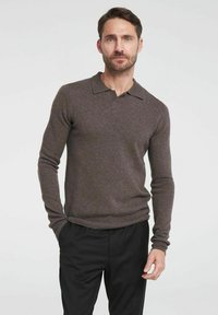 Bruine gebreide trui met een polokraag en lange mouwen, met een geribbelde zoom. Draagt met zwarte broek tegen een effen achtergrond.