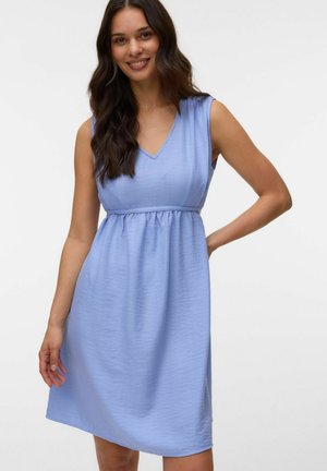 Femme souriante aux longs cheveux foncés portant une robe sans manches bleu clair longueur genou avec un col en V, debout devant un fond blanc uni.