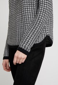 Schwarzer und weißer gemusterter Strickpullover mit strukturierter Oberfläche, rundem Ausschnitt und gerippten Bündchen, kombiniert mit schwarzen Stoffhosen.