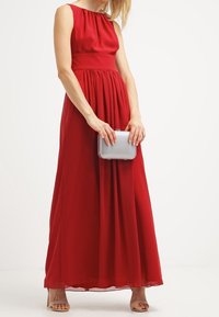 Donna in un elegante abito da sera senza maniche di colore rosso, che tiene in mano una piccola borsa a tracolla argentata, indossando tacchi aperti beige, in piedi su uno sfondo bianco.