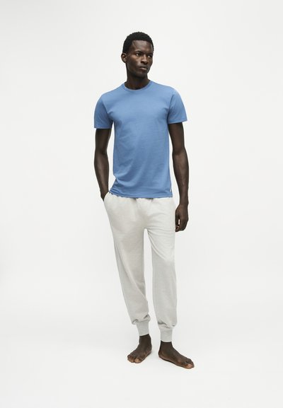 Jeune homme debout pieds nus, portant une chemise bleue à manches courtes et un pantalon de survêtement gris clair sur un fond blanc uni.